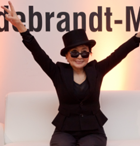 Yoko Ono sitzt lächelnd auf einer Couch und zeigt mit beiden Händen das Victory-Zeichen